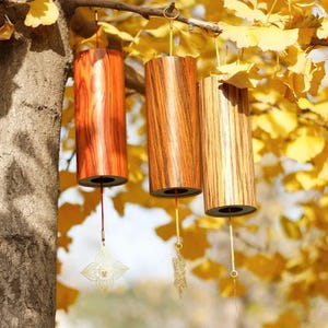 May include: Three wooden wind chimes with a cylindrical shape hang from a tree branch. The chimes have a natural wood grain finish in shades of brown and orange. Each chime has a decorative gold-colored charm hanging from a string.
