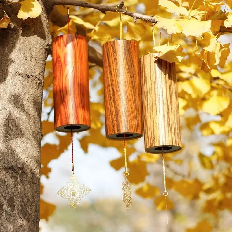 May include: Three wooden wind chimes with a cylindrical shape hang from a tree branch. The chimes have a natural wood grain finish in shades of brown and orange. Each chime has a decorative gold-colored charm hanging from a string.