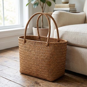 May include: A rectangular woven basket with two braided handles. The basket is made of a light brown material and has a textured surface. The basket is on a wooden floor.