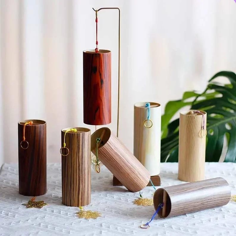 May include: A collection of cylindrical wooden wind chimes in various wood tones, including dark brown, light brown, and reddish-brown. Some chimes are upright, while others are angled or lying down. Each chime has a small ring and a colorful string.