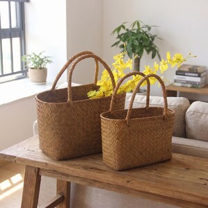 May include: Two woven brown baskets with handles sit on a wooden table. The larger basket is filled with yellow flowers. The baskets have a rectangular shape and are made of a natural fibre material.