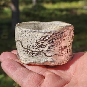 Puede incluir: Una pequeña taza cilíndrica de cerámica con un acabado de esmalte blanquecino agrietado. El exterior presenta una ilustración dibujada a mano en marrón oscuro de un dragón con líneas fluidas y serpentinas sobre la superficie.