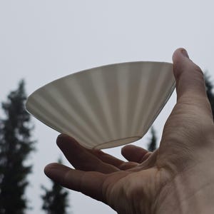 Puede incluir: Un cuenco de cerámica blanco acanalado sostenido en una mano contra un fondo de árboles y un cielo nublado. El cuenco tiene forma cónica con estrías verticales, creando una superficie texturizada. El cuenco está vacío y parece limpio.