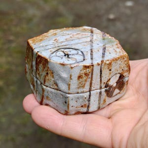 以下が含まれることがあります： 茶色の酸化鉄の斑点と濃い茶色の縦線が入った、質感のあるオフホワイトの釉薬が施された六角形の陶器製蓋付きボックス。蓋には手描きの円形の植物モチーフが描かれており、側面には幾何学模様があしらわれています。