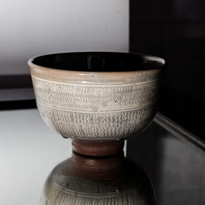 May include: A ceramic bowl with a dark interior and a light beige exterior. The bowl features a textured, wave-like pattern around the sides and sits on a brown pedestal base. The bowl is displayed on a reflective surface.