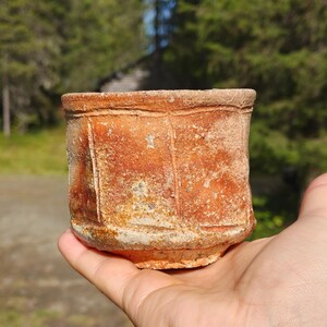 Può includere: Una piccola tazza cilindrica in ceramica con una superficie ruvida e strutturata in tonalità arancione bruciato e marrone terra. L'esterno presenta rientranze verticali che creano un effetto a pannelli, con una finitura in argilla granulosa.