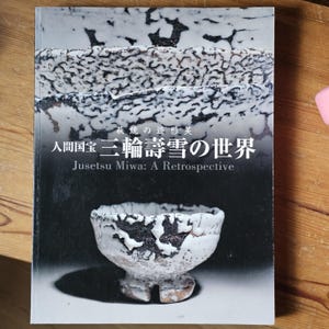 Puede incluir: Una portada de libro con cuencos de cerámica blancos y negros. El título es "Jusetsu Miwa: A Retrospective" en inglés, con texto japonés encima. El libro está sobre una superficie de madera.