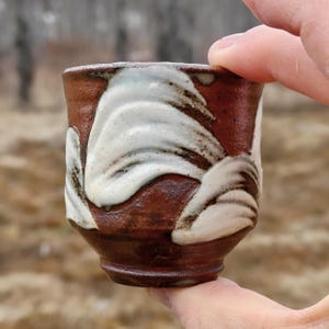 May include: A handmade ceramic cup with a brown base and white, wave-like patterns. The cup is held by a person's fingers, showcasing its unique design and texture. The cup is likely used for drinking beverages.