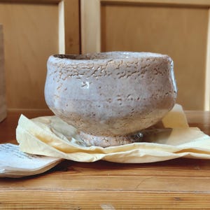 May include: A handcrafted, off-white ceramic bowl with a textured surface. The bowl sits on a yellow cloth, with a wooden background. The bowl has a wide, open top and a slightly rough, organic texture.