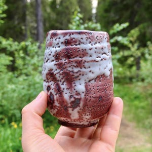 May include: A cylindrical ceramic cup featuring a textured, cratered surface with a dual-tone glaze. The exterior displays a deep reddish-brown base color partially overlaid with a thick, drippy white glaze that creates a mottled, porous appearance.