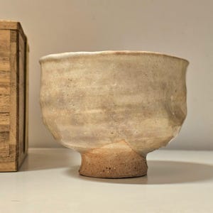 Puede incluir: Un cuenco de cerámica blanco roto hecho a mano, con una superficie texturizada y una base pequeña y redondeada. El cuenco tiene una abertura ancha y una forma ligeramente irregular. Hay una caja de madera al lado.