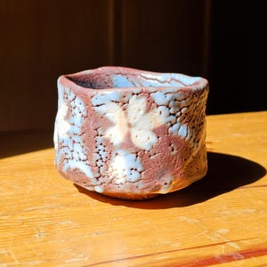 May include: A handmade ceramic cup with a textured surface. The cup features a blend of colors, including brown, blue, and white, creating a unique, organic appearance. The cup is sitting on a wooden surface.