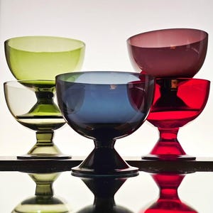 Op de afbeelding: Een verzameling kleurrijke glazen dessertkommen in groen, blauw, rood en paars. Elke kom heeft een afgeronde vorm en staat op een korte, steelachtige voet. De kommen zijn gerangschikt op een reflecterend oppervlak.