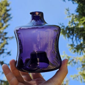 Puede incluir: Un jarrón de cristal morado oscuro con un cuerpo acampanado y un cuello estrecho. El jarrón se sostiene contra un fondo de cielo azul y árboles verdes. El texto "MADE IN FINLAND" es visible en la parte superior del jarrón.