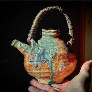 Pode incluir: Uma chaleira de cerâmica artesanal com uma alça trançada. A chaleira apresenta uma superfície texturizada com uma mistura de tons laranja, castanho e verde. O bico e a alça estão intactos, mostrando a habilidade do artesão.