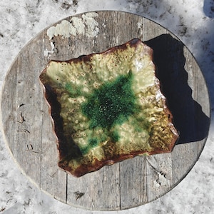 May include: A square ceramic plate with a unique, textured design. The plate features a central area of deep green, surrounded by a mottled, light green and brown border. The edges have a rough, uneven appearance, adding to its handcrafted aesthetic.