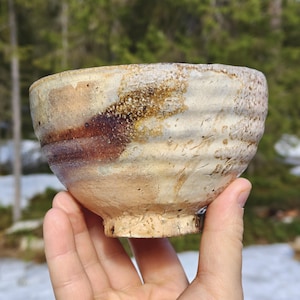 Può includere: Una ciotola in ceramica fatta a mano con una superficie strutturata. La ciotola presenta una palette di colori beige e marrone con un'estetica rustica e organica. Il design include una base a piede e un bordo leggermente irregolare.