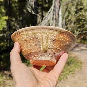 May include: A handcrafted ceramic bowl with a speckled, rustic finish. The bowl displays a warm colour palette of brown, orange, and cream, with subtle green accents. The bowl is held in a hand, highlighting its unique shape and texture, ideal for serving food.