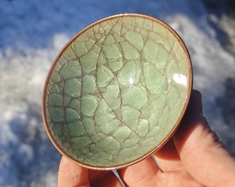 May include: A small, oval-shaped ceramic bowl with a celadon green glaze and a crackle pattern. The rim has a copper-colored trim. The bowl is held in a hand, with a snowy background.