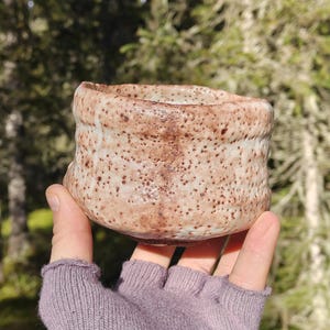 Könnte beinhalten: Eine kleine, runde Keramiktasse mit einer texturierten, gesprenkelten Oberfläche in hellen Beige-, Braun- und Erdtönen. Das Gefäß hat einen unebenen, organischen Rand und eine gesprenkelte Glasur mit dunkelbraunen Punkten auf der Außenseite.