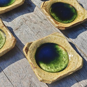 May include: Four handmade ceramic dishes with a square shape and a circular, glazed center. The centers are a mix of blue and green, while the outer edges are a tan color. The dishes are arranged on a weathered wooden surface.
