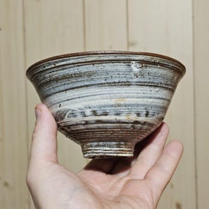 Op de afbeelding: Een handgemaakte keramische kom met een uniek, getextureerd ontwerp. De kom heeft een bruine rand en basis, met een wervelend patroon van witte en bruine tinten aan de buitenkant. De kom wordt in een hand gehouden.