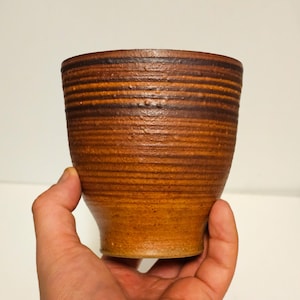 May include: A handmade ceramic cup with a textured, horizontal striped design. The cup features a warm, earthy color palette of brown and tan. The cup is held in a hand against a white background.