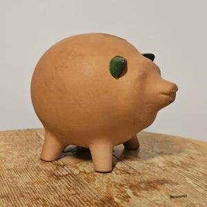 May include: A terracotta-colored piggy bank with green accents on the ears. The ceramic bank has a rounded body, short legs, and a small snout. The piggy bank is sitting on a wooden surface.
