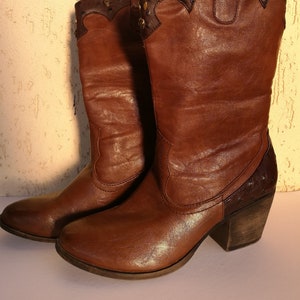 Puede incluir: Un par de botas vaqueras de cuero marrón con un detalle de costura decorativo. Las botas tienen una punta puntiaguda y un tacón de bloque.