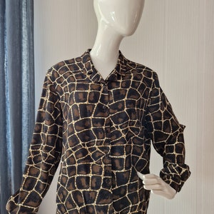 Puede incluir: Una camisa de manga larga con botones y estampado de animal marrón y dorado. La camisa tiene cuello y un corte holgado.