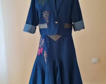 May include: A denim skirt and jacket set with a dark blue color. The skirt has a flared shape and the jacket has a cropped style. Both pieces feature embroidered floral designs with beaded accents.