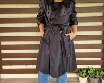 May include: A black trench coat with a double-breasted design, a belt, and pockets. The coat is knee-length and has long sleeves. The person is wearing blue jeans and leopard print shoes. The background features a wooden slatted wall and a red rug.