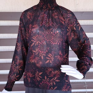 May include: A black sheer blouse with a floral print in shades of red and orange. The blouse has a high neck and long sleeves with elastic cuffs.