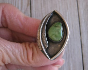 May include: A marquise-shaped ring featuring a polished green stone set in oxidized silver. The metal frame displays a detailed feather motif along one side, with a smooth, dark, and textured surface surrounding the central stone setting.