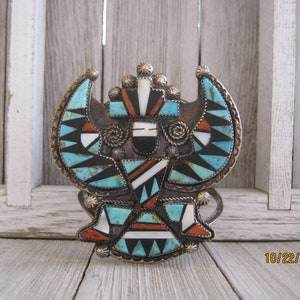 May include: A silver and turquoise Native American style belt buckle with intricate geometric designs. The buckle is made of silver and features a variety of turquoise stones, as well as other stones in black, white, and red.