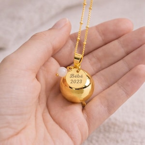 May include: A gold-colored necklace with a round pendant that has the text "B&eacute;b&eacute; 2023". The necklace has a small white bead and a gold chain. The necklace is held in a hand.