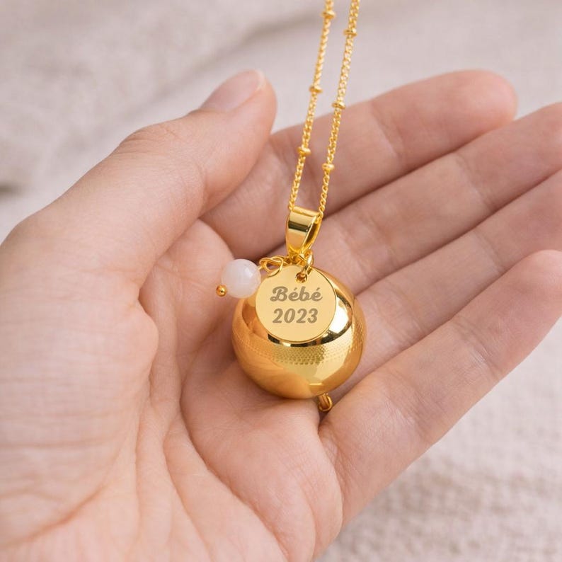 May include: A gold-colored necklace with a round pendant that has the text "B&eacute;b&eacute; 2023". The necklace has a small white bead and a gold chain. The necklace is held in a hand.