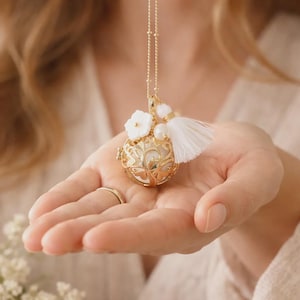 Peut inclure: Un collier pendentif doré avec un motif en filigrane, des fleurs blanches et un pompon blanc. Le collier est tenu dans une main ouverte. Le pendentif mesure environ 2,5 cm de diamètre.