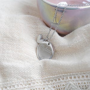 May include: A silver-colored necklace with a heart-shaped charm and a round pendant. The pendant is engraved with the name "Mylann" in cursive script. The necklace is displayed on a cream-colored fabric.