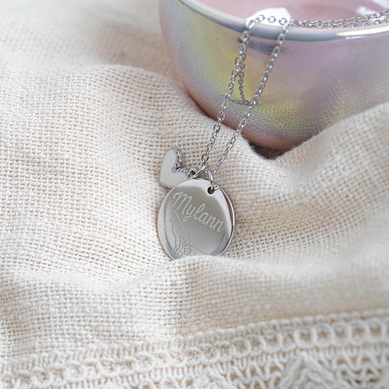 May include: A silver-colored necklace with a heart-shaped charm and a round pendant. The pendant is engraved with the name "Mylann" in cursive script. The necklace is displayed on a cream-colored fabric.