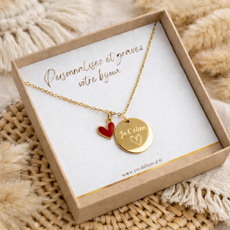 May include: A gold-colored necklace with a heart-shaped pendant and a round pendant. The round pendant has the words "Je t'aime" and a heart symbol engraved on it. The necklace is presented in a cardboard box with text that reads "Personnalisez et gravez votre bijoux."