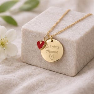 May include: A gold-colored necklace with a round pendant and a small red heart charm. The pendant is engraved with the words "Je t'aime Mamie" and a heart symbol. The necklace is displayed on a white surface.