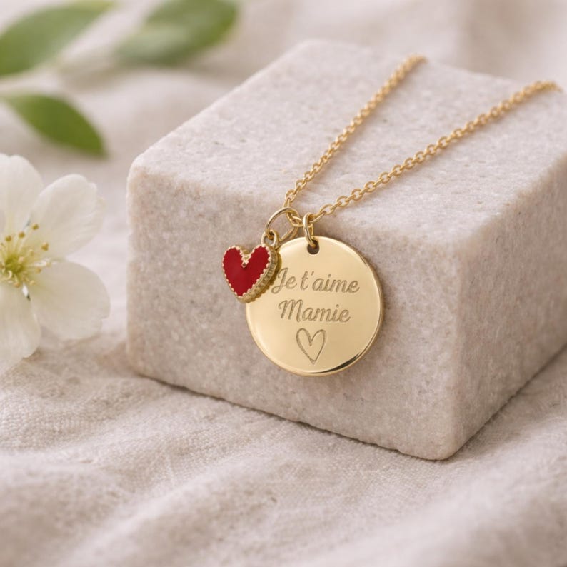 May include: A gold-colored necklace with a round pendant and a small red heart charm. The pendant is engraved with the words "Je t'aime Mamie" and a heart symbol. The necklace is displayed on a white surface.