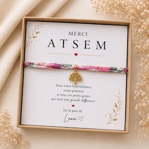 Könnte beinhalten: Ein Stoffarmband mit rosa-grünem Blumenmuster und einem goldenen Baum-Anhänger, präsentiert in einer Pappschachtel auf einer Karte mit dem Text MERCI ATSEM und einer Dankesbotschaft von Lana, verziert mit kleinen roten Herzen.