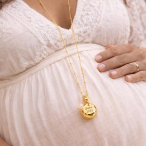 May include: A gold-colored necklace with a round pendant that says "Babe 2023". The necklace is long and delicate, with small beads along the chain. The pendant has a pearl-like accent. The person is wearing a white dress.