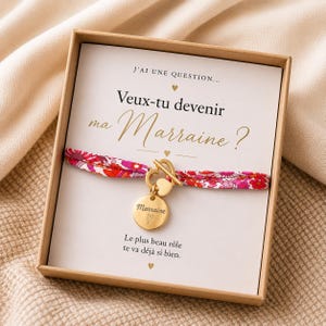 Op de afbeelding: Een goudkleurige toggle-armband met een bloemrijk stoffen bandje in roze en rode tinten, voorzien van een ronde bedel gegraveerd met het woord Marraine en een hartje. De armband zit in een vierkante kartonnen doos met een bedrukte kaart.