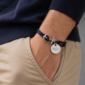Puede incluir: Pulsera azul marino con cierre de ancla plateado y un charm redondo grabado con el nombre "Mahé". La pulsera está hecha de un material similar a una cuerda azul oscuro.