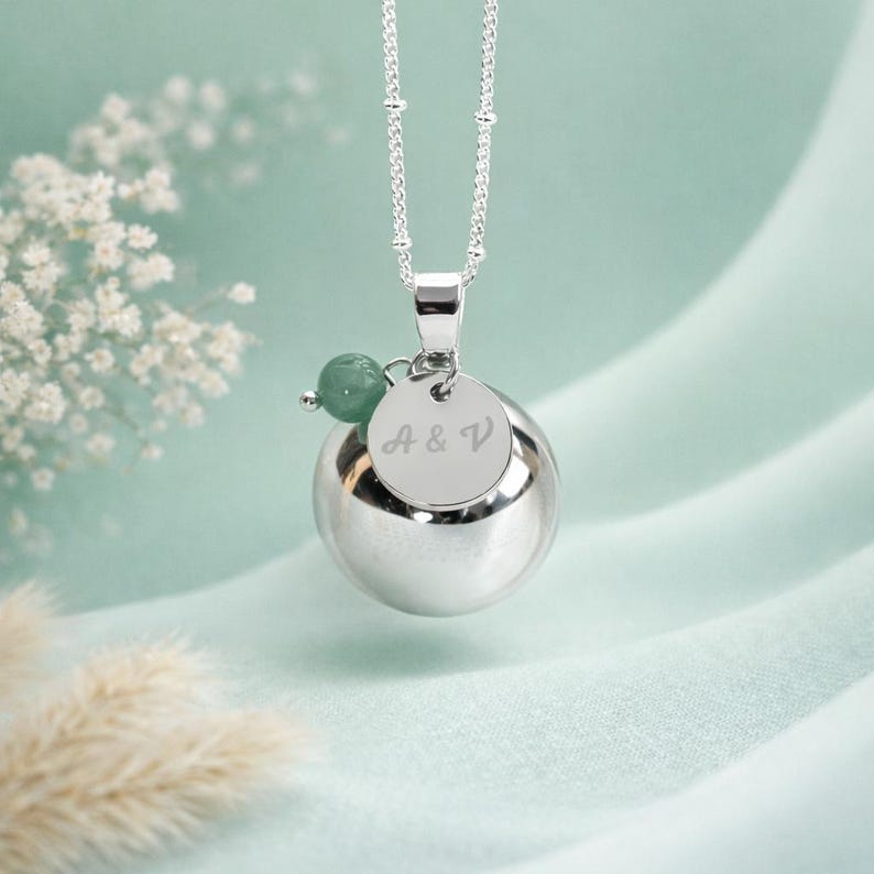 May include: A silver necklace with a round pendant and a small green bead. The pendant has the engraved initials "A & V" on a small, round disc. The necklace hangs on a delicate chain against a soft, blurred background.