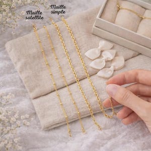 May include: Gold-colored necklaces in various chain styles, including a satellite chain and a simple chain, are displayed on a neutral-toned fabric. The image also shows a hand holding a necklace, along with small white bows and a jewelry box.