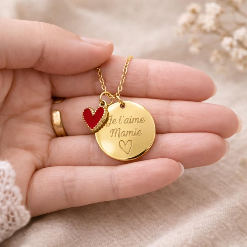 May include: Gold-colored necklace with a pendant featuring a red heart charm and a round disc. The disc is engraved with the words "Je t'aime Mamie" and a heart symbol. The necklace is a delicate piece of jewelry.